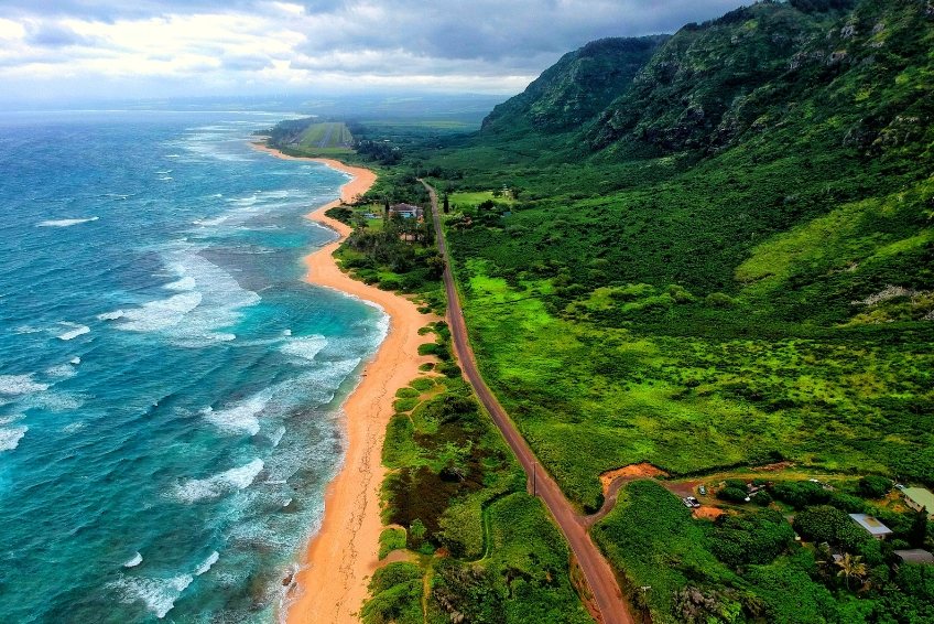 Aerial view of Oahu, Hawaii showing dramatic green mountain cliffs, turquoise ocean with white waves, golden sand beach, and coastal road stretching along the shoreline