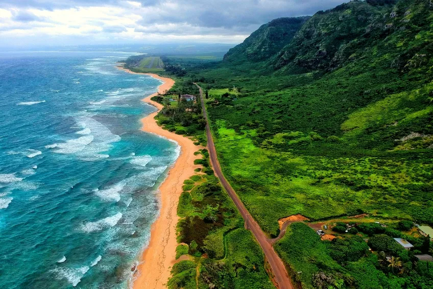 Aerial view of Oahu, Hawaii showing dramatic green mountain cliffs, turquoise ocean with white waves, golden sand beach, and coastal road stretching along the shoreline